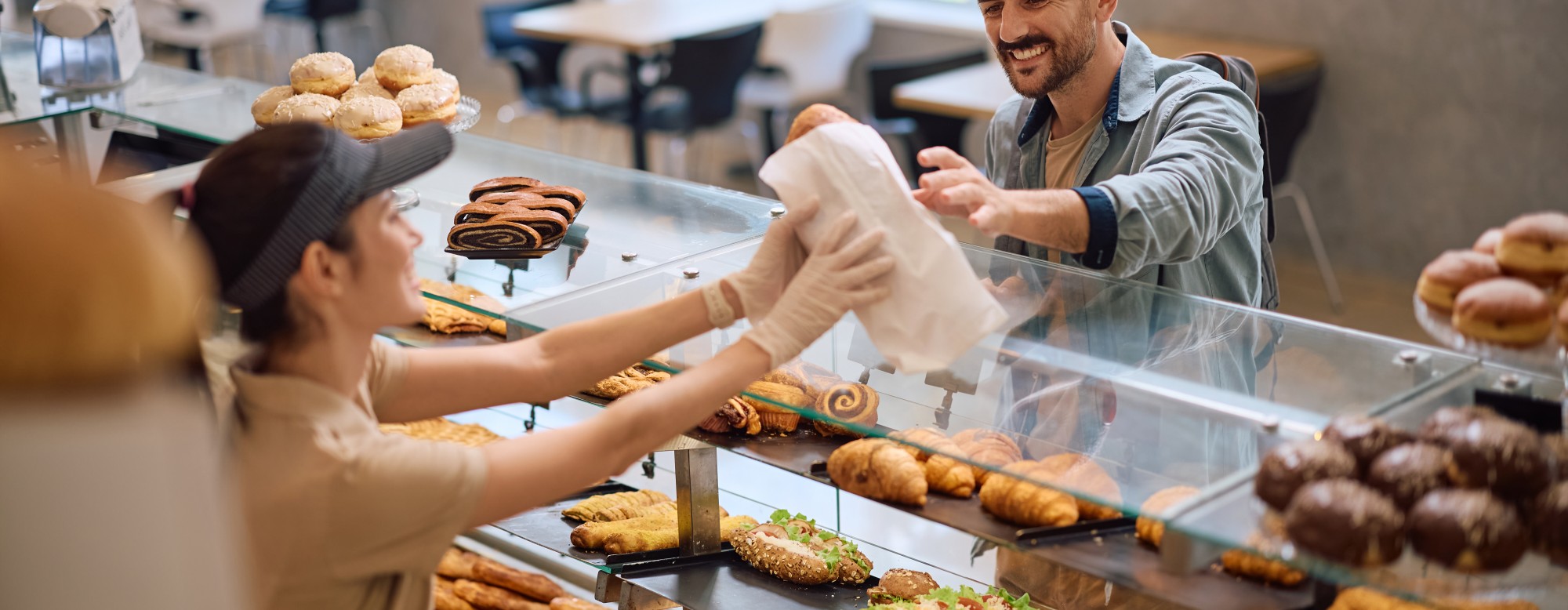 Un client souriant reçoit un sac de viennoiseries d’une vendeuse dans une boulangerie moderne.